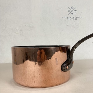 Könnte beinhalten: Ein runder Kupfertopf mit einem dunklen Metallgriff, der mit zwei Kupfernieten befestigt ist. An der Seite des Topfes befindet sich ein Stempel mit der Aufschrift E. DEHILLERIN 18 RUE COQUILLIERE. Die Außenfläche ist poliert und glänzend.