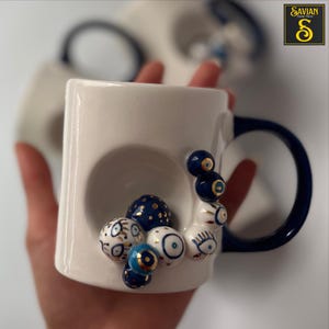 May include: White ceramic mug with a blue handle and a design of blue and gold evil eyes. The mug has a unique shape with a recessed area on the side.