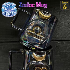 Könnte beinhalten: Zwei schillernde Zodiac-Tassen aus Keramik, die übereinander gestapelt sind. Die Tassen haben einen schwarzen Griff und ein goldenes Waage-Symbol. Der Text "Zodiac Mug" steht über den Tassen, und das Waage-Symbol befindet sich links.