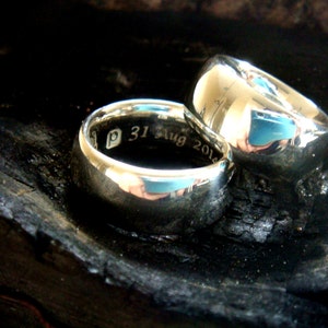 May include: Two silver wedding bands, one with the inscription "P 31 Aug 2014", resting on a dark, textured surface.