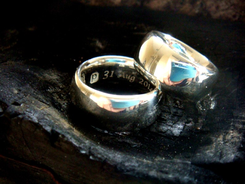 May include: Two silver wedding bands, one with the inscription "P 31 Aug 2014", resting on a dark, textured surface.