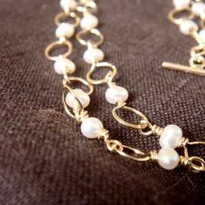 May include: A gold chain necklace with white pearl beads. The chain is made of two strands of gold wire, each with a series of small, round loops. The pearls are evenly spaced along the chain.