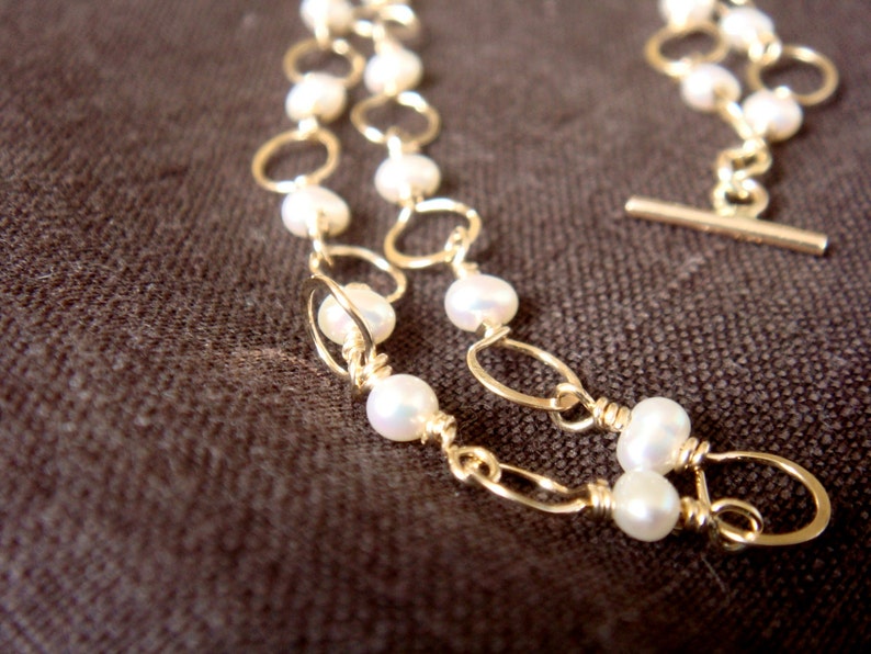 May include: A gold chain necklace with white pearl beads. The chain is made of two strands of gold wire, each with a series of small, round loops. The pearls are evenly spaced along the chain.