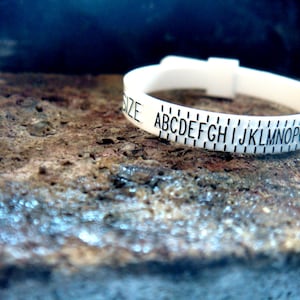 Peut inclure: Anneau en plastique blanc avec inscription noire "SIZE ABCDEFGHIJKLMNOPQRSTUVWX".