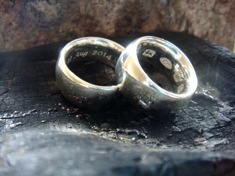 May include: Two silver wedding bands with a rounded profile. The band on the left has the inscription "1 Aug 2014". The band on the right has the inscription "925".
