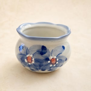 May include: A small, round ceramic planter with a scalloped rim. The exterior is white with hand-painted blue floral motifs featuring dark blue petals and orange centers surrounded by small white dots. The rim is accented with a light blue wash.