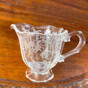 May include: Clear glass creamer with a handle and a floral pattern etched into the glass. The creamer has a scalloped rim and sits on a pedestal base. The creamer is designed for serving cream or milk.