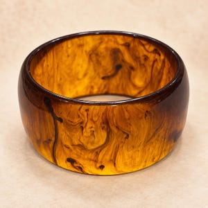May include: A wide, circular bangle bracelet featuring a translucent amber-colored base with dark brown, swirling, smoke-like patterns throughout the material. The surface is smooth and polished, creating a continuous, rounded band design.