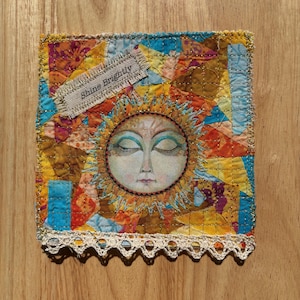 May include: A colorful textile art piece featuring a sun face with closed eyes. The artwork has a patchwork background in shades of orange, blue, and gold. The text "Shine Brightly" is on a small label. A white lace trim adorns the bottom edge.
