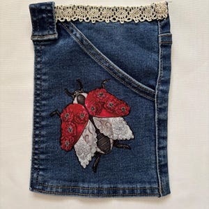 May include: A blue denim pocket featuring a decorative illustration of a beetle. The beetle has red wings adorned with floral patterns and white lace-like wing details. The top edge of the denim pocket is trimmed with a cream-colored crochet lace border.