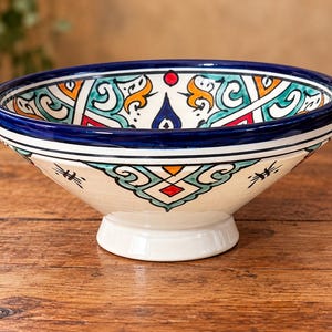May include: A ceramic pedestal bowl featuring a cream-colored base with hand-painted geometric and floral patterns in shades of deep blue, turquoise, orange, and red. The bowl has a thick dark blue rim and a flared footed base.