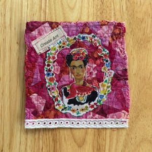 May include: A quilted textile art piece featuring a portrait of Frida Kahlo within a floral frame. The artwork is predominantly shades of pink and red, with a white lace trim at the bottom. The text "Create Art" is on a small tag.