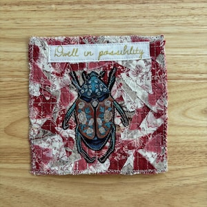 May include: A square, quilted fabric piece with a red and white floral pattern. A detailed illustration of a blue and brown beetle with floral accents is centered on the fabric. The text "Dwell in possibility" is embroidered in gold.