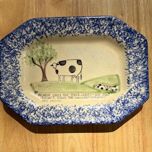 May include: A rectangular ceramic serving platter with a blue and white sponge-painted border. The center features a hand-painted scene of a black and white cow near a tree, with text reading: MICHELE LIKED THE OTHER LADIES-SHE JUST COULDN'T STAND THE CONSTANT CHEWING AND MOOING.