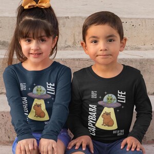 Puede incluir: Dos niños con camisetas de manga larga con un diseño de dibujos animados. El diseño presenta un carpincho con gafas de sol debajo de un OVNI con un extraterrestre, el texto "CAPYBARA LIFE" y "OUT OF THIS WORLD". Una camiseta es azul marino, la otra negra.