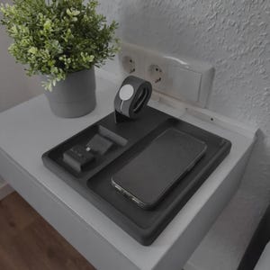 Peut inclure: Un plateau de chargement rectangulaire gris foncé doté de compartiments encastrés dédiés pour un smartphone, une montre connectée et des écouteurs sans fil. Le plateau présente une finition mate et offre un système d'organisation structuré.