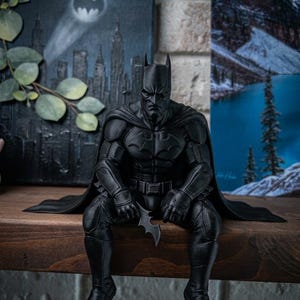 Könnte beinhalten: Eine mattschwarze Batman-Figur in sitzender Position, die einen detaillierten gepanzerten Anzug mit Umhang trägt und ein kleines fledermausförmiges Emblem in einer Hand hält. Die Figur zeigt eine modellierte Maske, Brustplatte und einen Gürtel.
