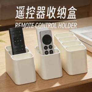 Peut inclure: Trois supports de télécommande en plastique blanc cassé avec plusieurs compartiments. Les supports présentent le texte "遥控器收纳盒" et "REMOTE CONTROL HOLDER" au-dessus. Deux supports contiennent des télécommandes noire et argentée.
