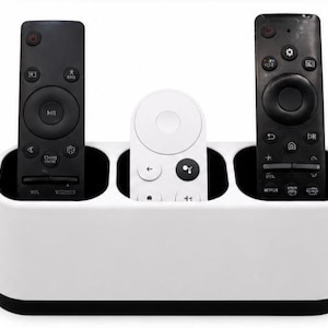 Peut inclure: Un support de télécommande rectangulaire blanc doté de trois compartiments distincts. Deux télécommandes noires sont placées dans les fentes extérieures, tandis qu'une télécommande blanche est positionnée dans la fente centrale.