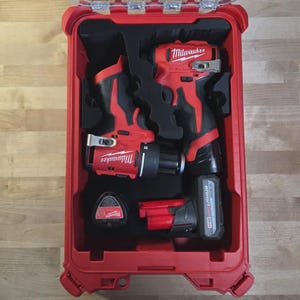 Puede incluir: Un maletín de transporte rígido rojo de Milwaukee que contiene un taladro inalámbrico rojo y negro, un atornillador de impacto rojo y negro, una batería y un pequeño accesorio circular, organizados en inserciones de espuma negra a medida.