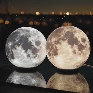 Peut inclure: Deux lampes sphériques en forme de lune posées sur une surface réfléchissante. La lampe de gauche émet une lumière blanche froide, tandis que celle de droite émet une lumière jaune chaude. Les deux présentent des textures détaillées de cratères.