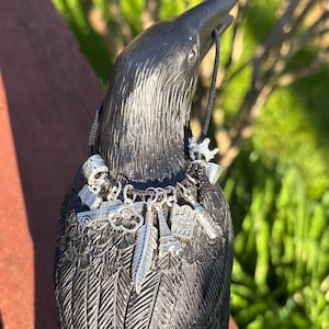 May include: A black raven figurine with a necklace of silver charms. The charms include a feather, a car, a skull, and a small Eiffel Tower. The raven is perched on a wooden surface.