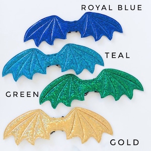 May include: Four pairs of glittery dragon wings in different colors: royal blue, teal, green, and gold. The wings are made of a soft, sparkly fabric and have a black elastic band for attaching them to a costume.