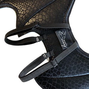 May include: Black dragon wings costume with a black, textured, scaly design. The wings have adjustable straps with buckles. The wings have a white label with the text "Mighty Fairy".