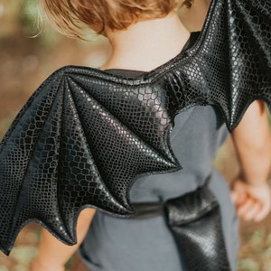 May include: Black dragon wings costume for kids. The wings are made of a soft, textured fabric with a scaly design. The wings are attached to a strap that goes around the shoulders.