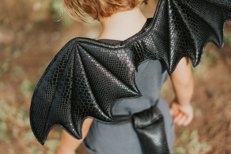 May include: Black dragon wings costume for kids. The wings are made of a soft, textured fabric with a scaly design. The wings are attached to a strap that goes around the shoulders.