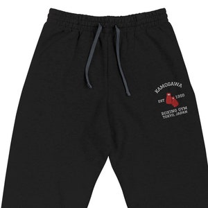 Puede incluir: Pantalones deportivos grises oscuros con cintura con cordón y puños acanalados. El texto "KAMOGAWA" está arqueado sobre un gráfico de guantes de boxeo rojos, con "EST. 1950" y "BOXING GYM TOKYO, JAPAN" impresos debajo.