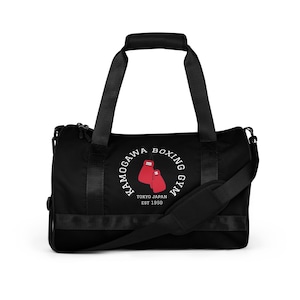 Peut inclure: Sac de sport noir avec un logo circulaire blanc représentant des gants de boxe rouges et le texte "KAMOGAWA BOXING GYM TOKYO JAPAN EST 1950". Le sac a une poignée supérieure, une bandoulière et une poche latérale.