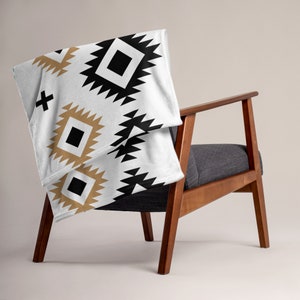 May include: A white throw blanket draped over a wooden chair. The blanket features a repeating geometric pattern in black and tan, with a black cross design. The chair has a dark gray seat and back.