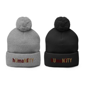 May include: Two beanies, one gray and one black, each with a matching pom-pom. The gray beanie has the word "hUMANiTY" embroidered in a reddish-brown color. The black beanie has the same text in the same color.