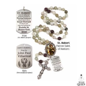 May include: A rosary with light green and brown beads, a silver cross, and a pendant with an image of St. Hubert, the patron saint of hunters. The pendant also has the text "St. Hubert" and a prayer.