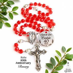 May include: A rosary featuring faceted red beads and white pearl-like beads on a silver-toned chain. It includes a silver crucifix, a centerpiece with a religious figure, and a round metal tag engraved with Jack & Liza Carlisle 40 Years April 26, 1986.