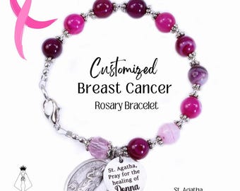 May include: A pink and purple beaded rosary bracelet with a silver clasp and a charm featuring St. Agatha, the patron saint of breast cancer. The charm has the text "St. Agatha, Pray for the healing of Donna".