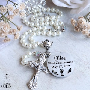 May include: A pearl rosary with a silver cross and a round tag that reads "Chloe First Communion May 17, 2025." The rosary is adorned with a silver chalice and iridescent beads. A silver tiara and floral accents are in the background.