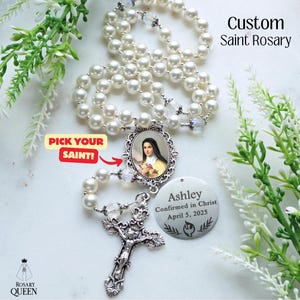 May include: A custom Saint Rosary with white faux pearls and a silver crucifix. The rosary features a saint pendant and a personalized tag. Text on the tag reads "Ashley Confirmed in Christ April 5, 2025". The image also includes the text "Pick Your Saint!"
