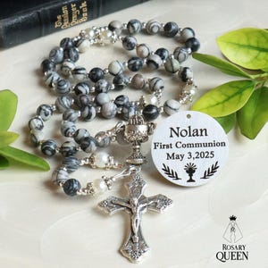 May include: A rosary with gray and white marbled beads, a silver crucifix, and a chalice. A white tag reads "Nolan First Communion May 3, 2025". The rosary is next to a book and green leaves.