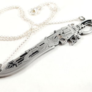 Puede incluir: Un collar plateado con un colgante de espada detallado. La espada tiene diseños intrincados y un mango negro.