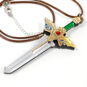 Può includere: Una collana con un ciondolo a forma di spada in argento e oro, con una gemma verde sul manico. Il ciondolo è attaccato a un cordino in pelle marrone.