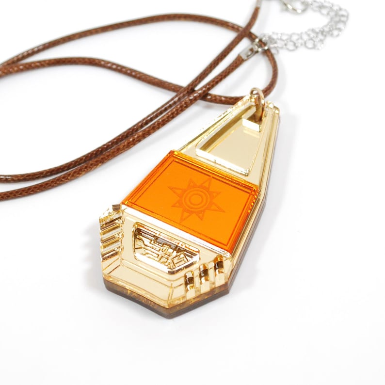 May include: A gold-colored pendant with a clear orange insert featuring a stylized sun design. The pendant is attached to a brown leather cord and a silver chain.