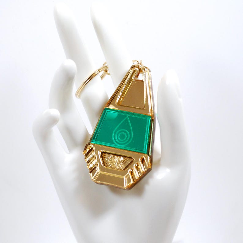 May include: Gold-colored keychain with a green, translucent, teardrop-shaped charm. The charm has a gold-colored frame and a stylized eye design.