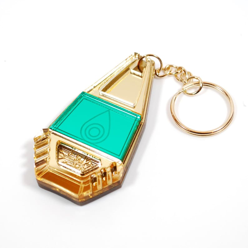 May include: Gold-colored keychain with a green, translucent, rectangular emblem featuring a stylized water drop with a circle inside. The keychain has a gold-colored ring.