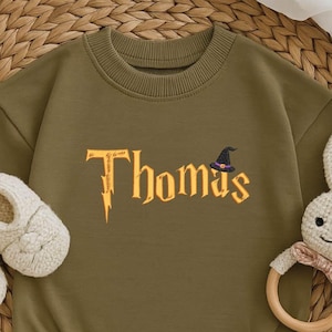 May include: Olive green baby romper with the name "Thomas" in gold lettering and a small witch hat graphic. The outfit is paired with cream-colored baby shoes and a stuffed bunny rattle. The romper has long sleeves and snap closures.