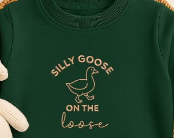 Op de afbeelding: Een donkergroene babybody met de tekst "Silly Goose on the Loose" in een speels lettertype. De body is omgeven door een rieten mand, een knuffel en een houten rammelaar. Het thema is babykleding.