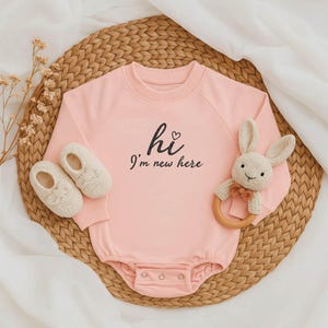 Könnte beinhalten: Ein hellrosa Baby-Strampler mit dem Text "hi I'm new here" in schwarzer Schrift. Der Strampler wird mit cremefarbenen Babyschuhen und einem Stoffhasen-Rassel mit Holzring kombiniert.