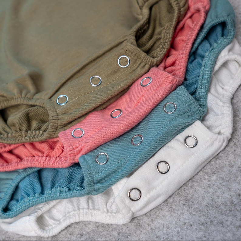 May include: Close-up of several baby rompers in various colors, including olive green, pink, teal, and white. Each romper has a row of silver snap closures. The fabric appears soft and comfortable.