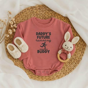 May include: A dusty rose baby onesie with the text "DADDY'S FUTURE running BUDDY" in black. The onesie is paired with white baby shoes and a bunny-shaped rattle with a wooden ring.